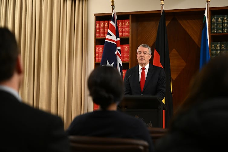 A man stands behind a lectern in front of flags