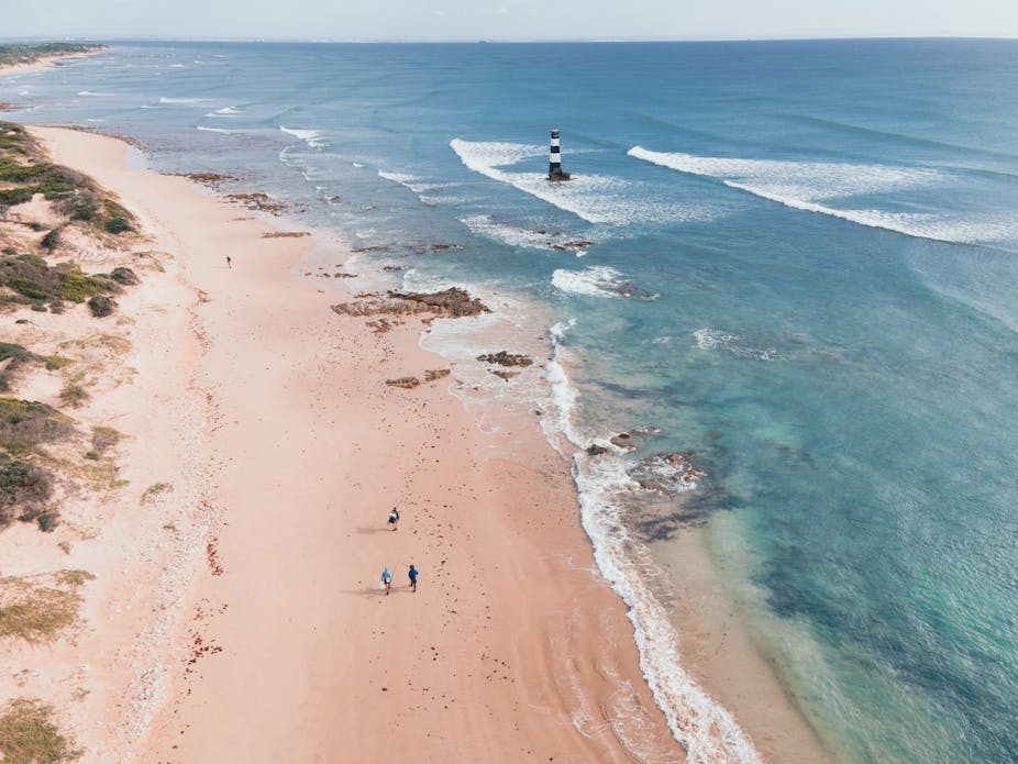 Aerial view of coastline with a lighthouse and people walking on the sandy beach.