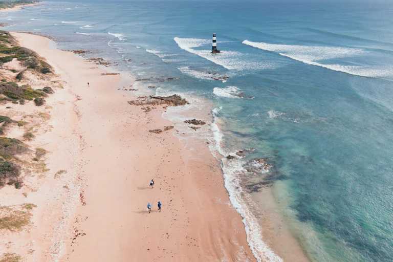 Aerial view of coastline with a lighthouse and people walking on the sandy beach.