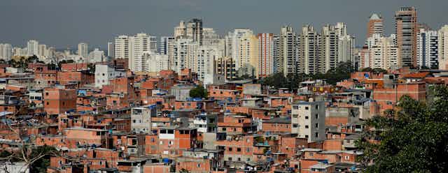 Vista de uma favela, com vários barracos de alvenaria, com prédios luxuosos ao fundo