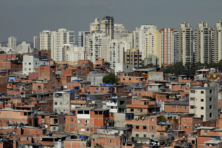 Vista de uma favela, com vários barracos de alvenaria, com prédios luxuosos ao fundo