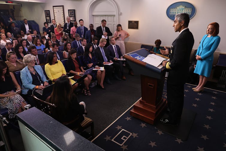 A man in a military uniform stands at a lectern looking out at a group of people sitting in chairs.