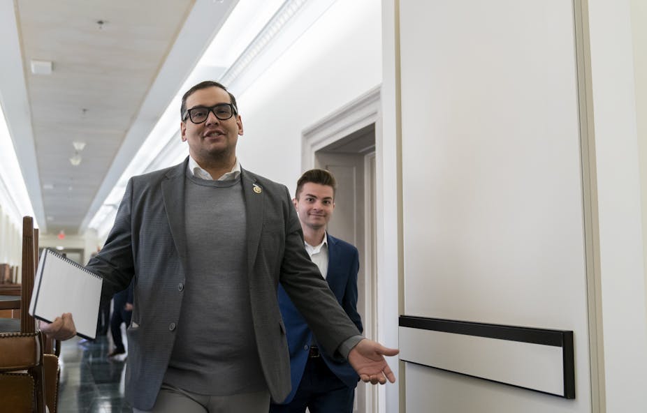 A bespectacled man, wearing a gray blazer, with a file in one hand, walking through a hallway, with another person right behind him.