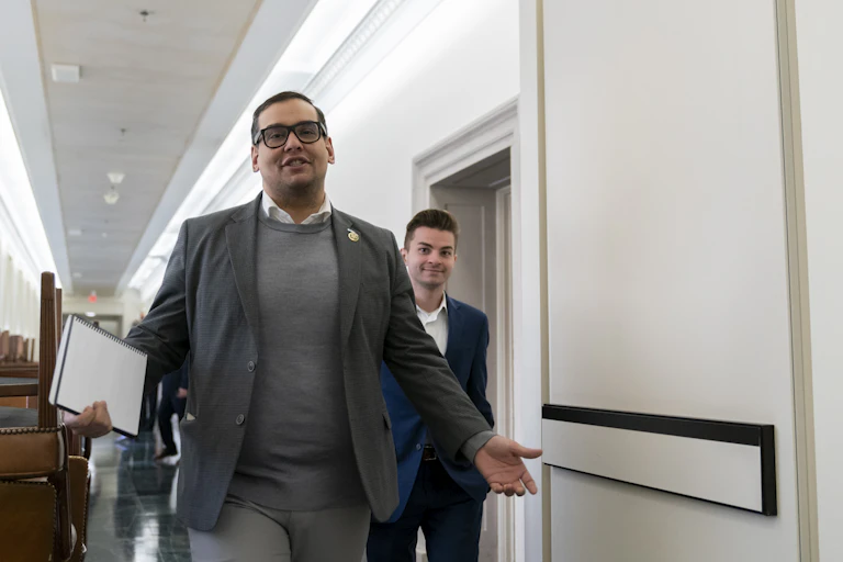 A bespectacled man, wearing a gray blazer, with a file in one hand, walking through a hallway, with another person right behind him.
