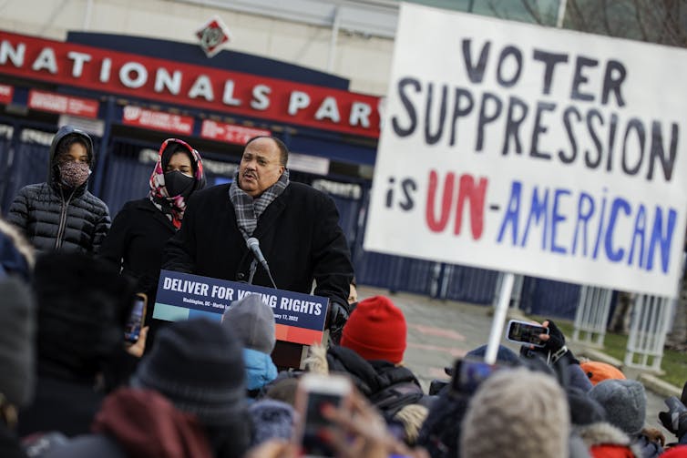 A Black middle-aged man speaks at a podium with the words, 'deliver for voting rights,' in a crowd of people who are wearing jackets. One person holds a sign that says 'voter suppression is un-American.'