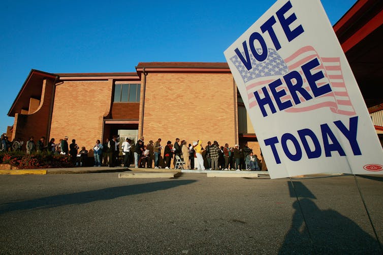 A big sign says 'vote here today' in front of a long line of Black people who stand ouside of a brown brick building.