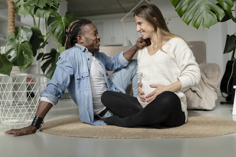 A man and a pregnant woman in casual clothing sit smiling on the floor as she holds her stomach.
