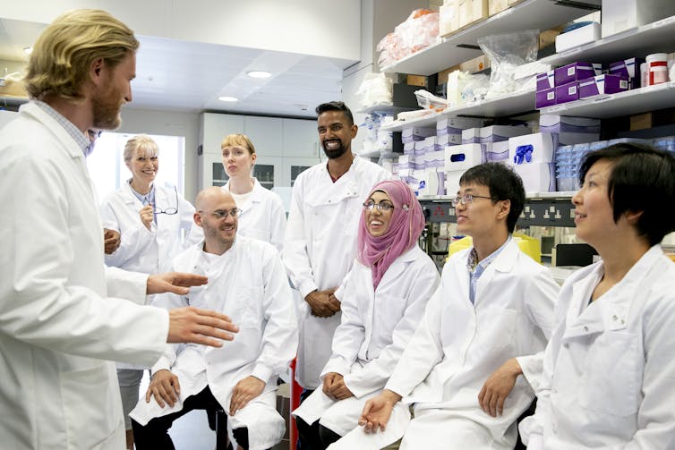A group of scientists in white coats sits laughing as they sit in an informal lab meeting.