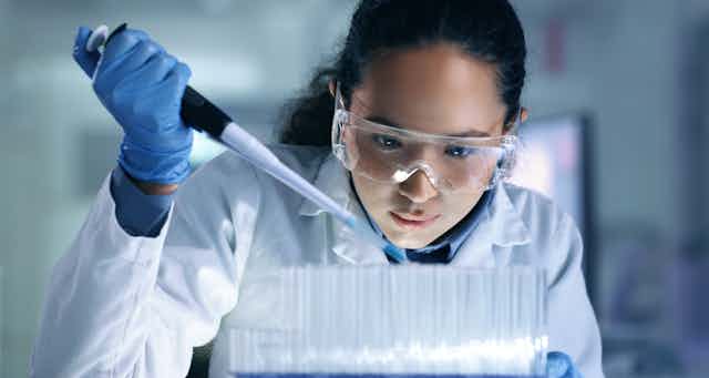 A young woman in a white lab coat, blue gloves and safety goggles inserts something into lab pipettes.