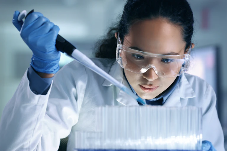 A young woman in a white lab coat, blue gloves and safety goggles inserts something into lab pipettes.