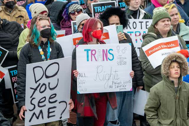 A group of young people, some with their hair dyed blue and pink, hold signs that say, 'Do you see me?' and 'Trans rights.'
