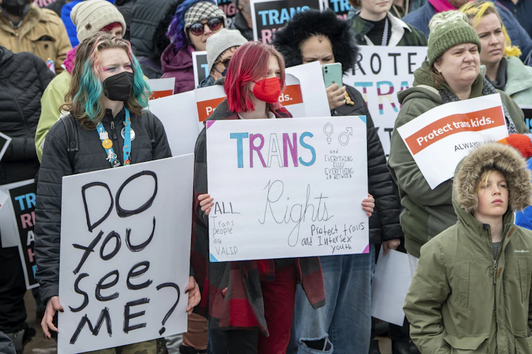 A group of young people, some with their hair dyed blue and pink, hold signs that say, 'Do you see me?' and 'Trans rights.'