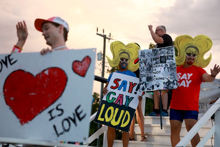 People hold signs that say 'Love is love,' and 'say gay loud!' Some of the people wear large yellow wigs.