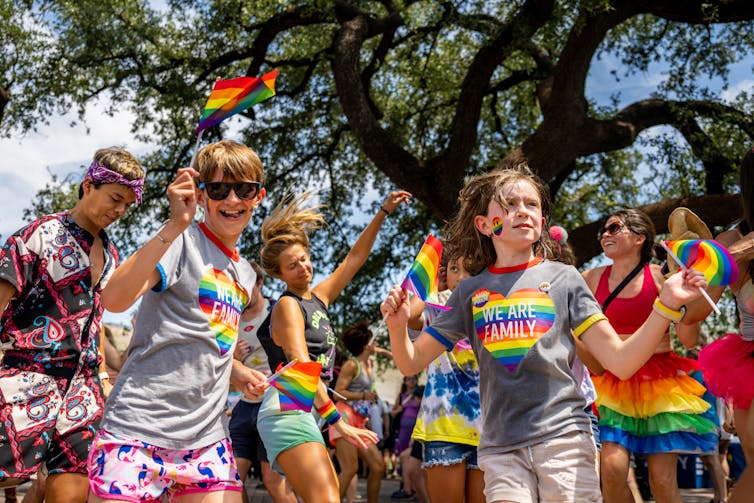 Los niños usan arcoíris en sus camisetas y bailan frente a personas que también usan arcoíris y ondean banderas de arcoíris.