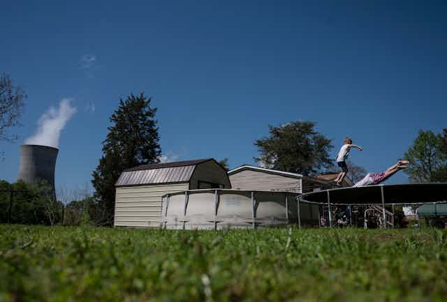 Two kids play on a trampoline next to an above-ground swimming pool. A power plant smokestack is in the background.