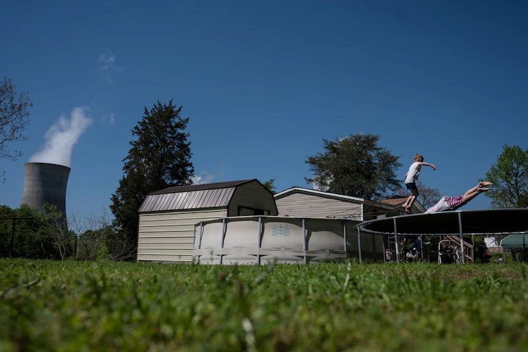 Two kids play on a trampoline next to an above-ground swimming pool. A power plant smokestack is in the background.