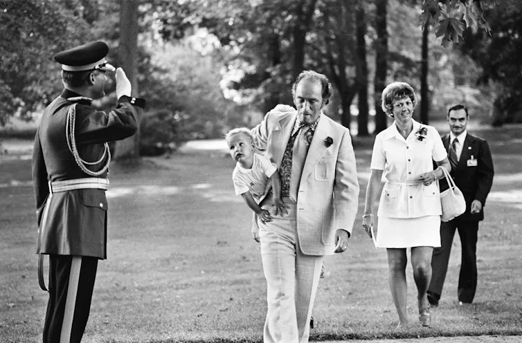 A black and white photo shows a man in a light suit with a rose in his lapel carrying a small boy who looks at an RCMP officer as he salutes his father.