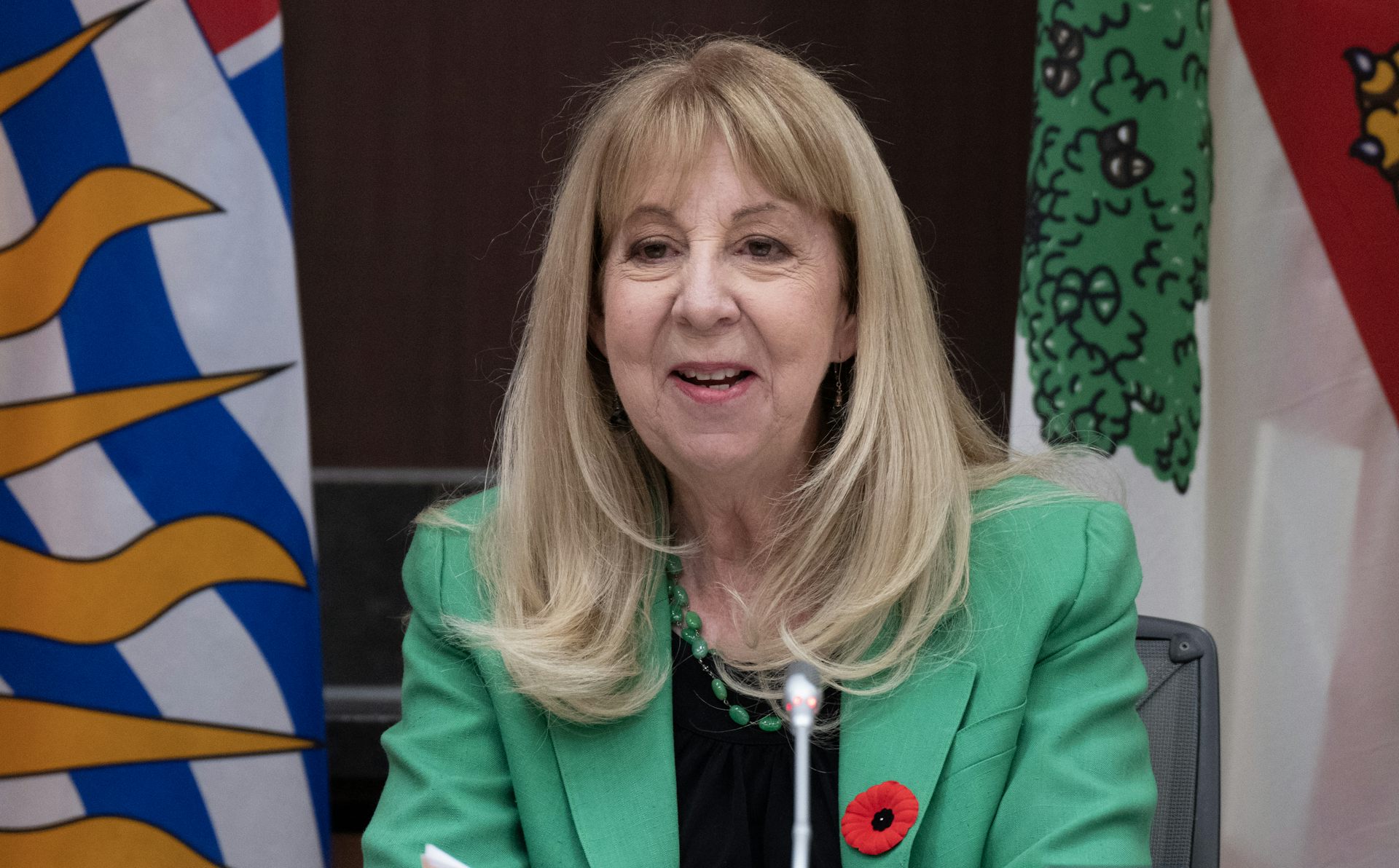 An older woman with shoulder-length blonde hair and a green jacket smiles while sitting behind a microphone.
