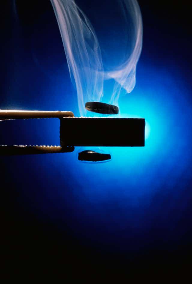 Two metal tongs holding a piece of black metal, with small metal discs hovering above and below it, with steam coming off and a light blue background. 