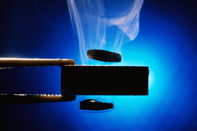 Two metal tongs holding a piece of black metal, with small metal discs hovering above and below it, with steam coming off and a light blue background.
