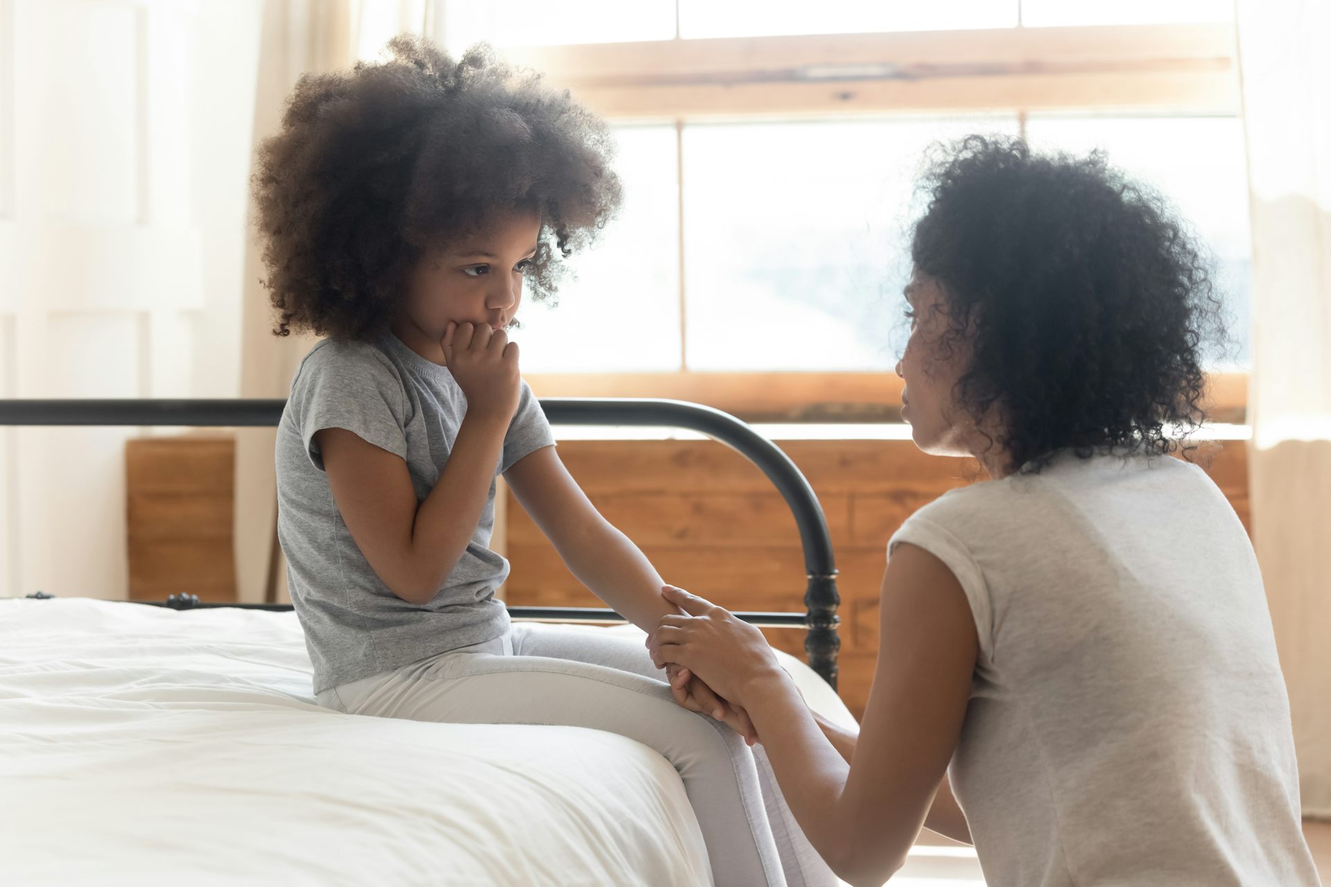 A black woman holds the hand of a young black girl sitting on a bed