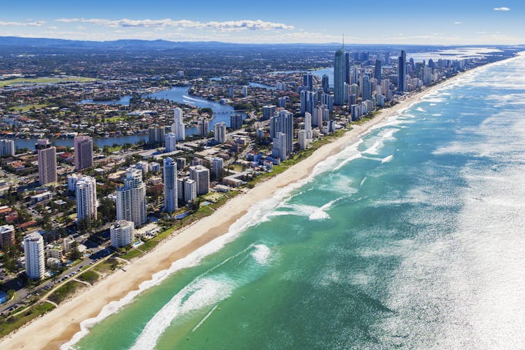 A high up view of an azure ocean coast right next to a highrise city with mountains in the background