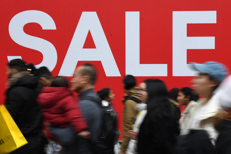 Pedestrians rushing past a large SALE sign on a shop window