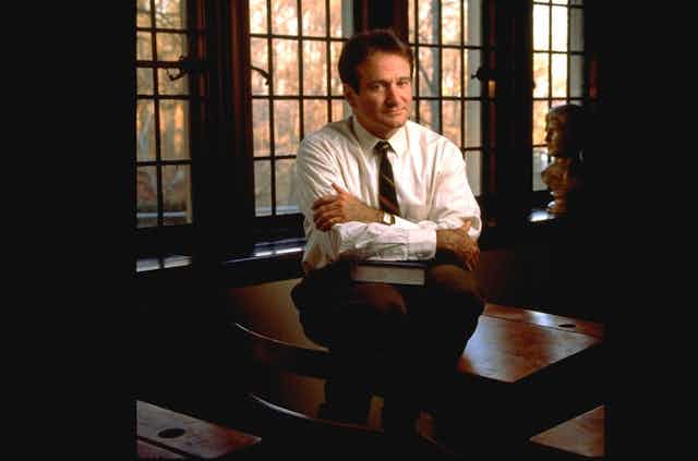 Actor Robin Williams sits on a desk in a still from the film Dead Poet's Society.
