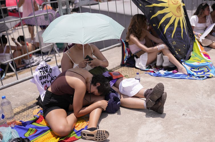 Taylor Swift fans wait outside stadium in extreme heat, under umbrellas.