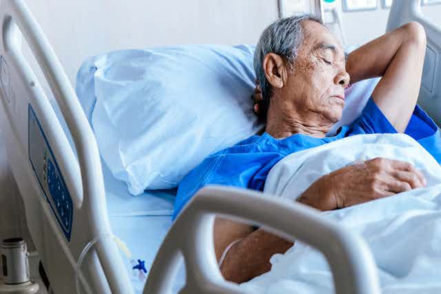 Older Asian man in hospital bed, resting or asleep, hand behind head