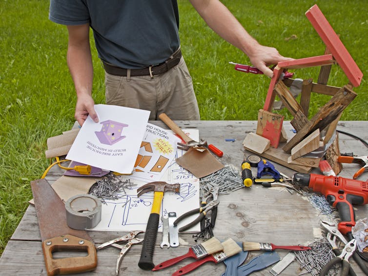 Person attempting to build a crooked bird house with tools strewn across a table