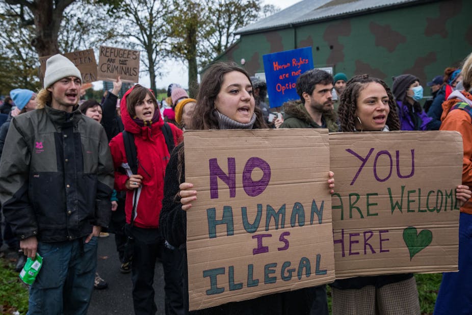 Men and women standing on the street, mouths open in a chant and holding up posters written 'No human is illegal', 'You are welcome here' and 'Refugees are not criminals'