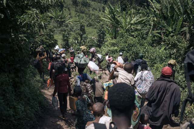 Men, women and children walking through a forested path with bags on their backs, heads and in their hands