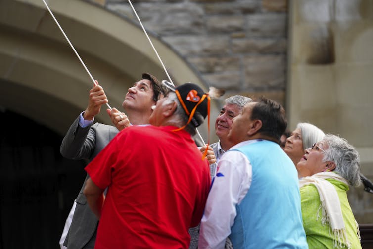 A dark-haired man and a group of older Indigenous people look up as they pull on a rope to raise a flag.