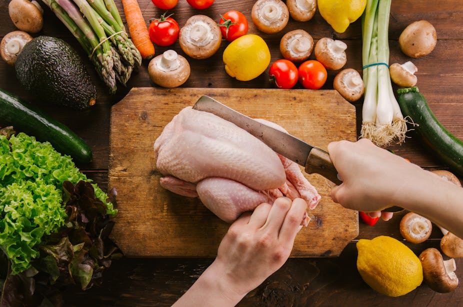 A person uses a butcher's knife to slice into a whole raw chicken on a cutting board.