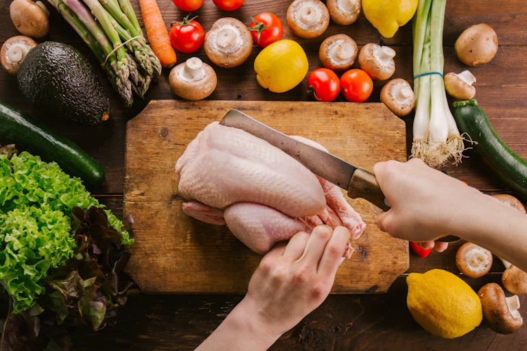 A person uses a butcher's knife to slice into a whole raw chicken on a cutting board.