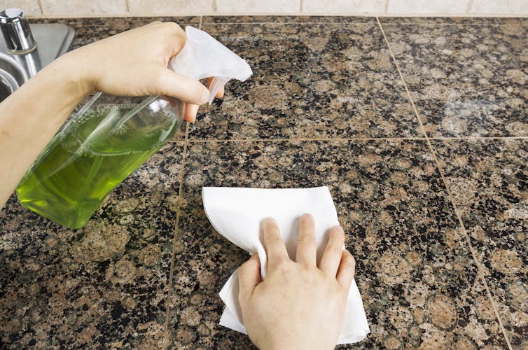 A person uses a spray bottle of kitchen disinfectant and a paper towel to clean a counter.