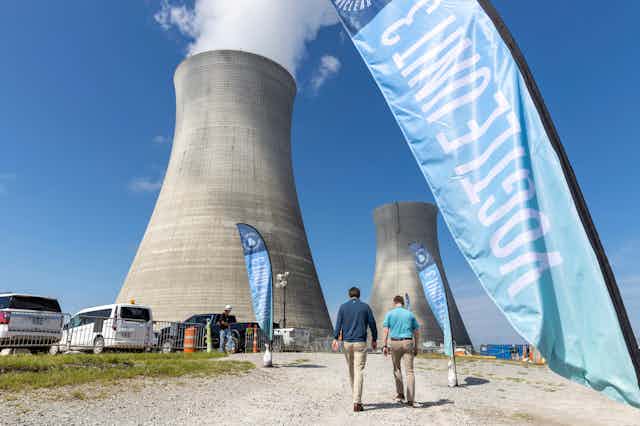 Two men walk towards a bank of cooling towers against a clear sky.