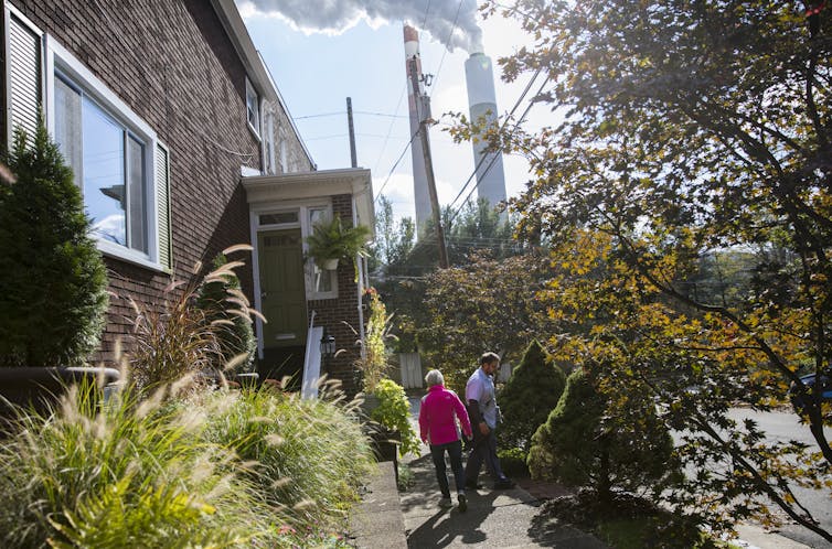 Two people stand outside an older brick home with power plant smokestacks in the background.