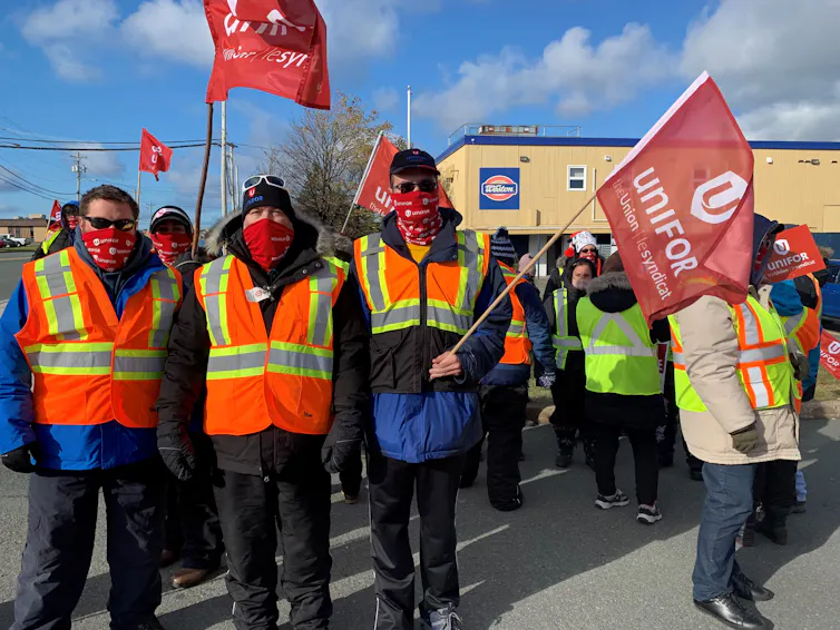 A group of people wearing coats and construction vests wave red flags that say UNIFOR on them