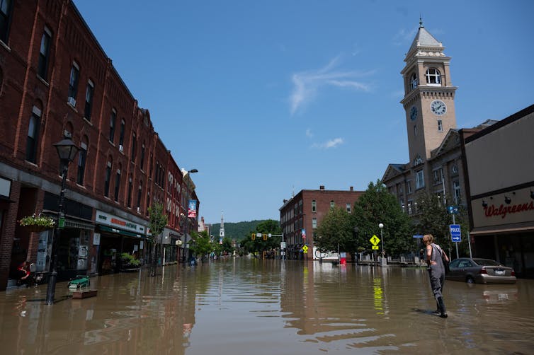 A person in high boots walks through water covering Main Street through a business district in Montpelier, Vermont. Water covers the sidewalks, streets and storefronts.