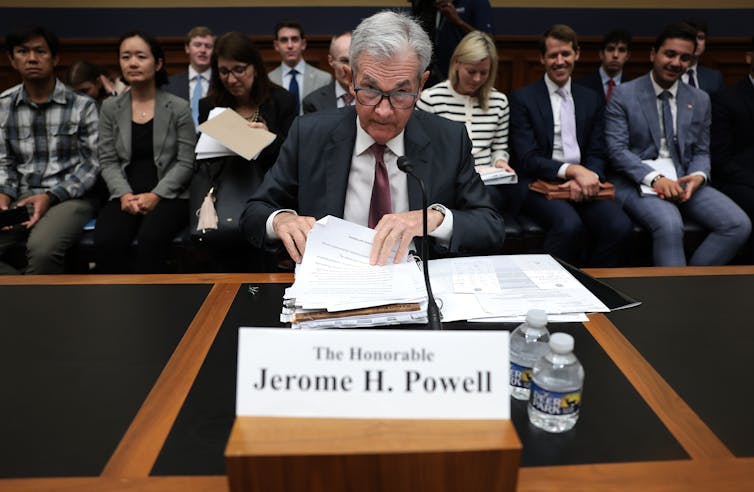 Powell, in a suit and tie, sits at a large desk in hearing room with papers in front of him and a name tag. He's  looking up over the top of his glasses at the camera.