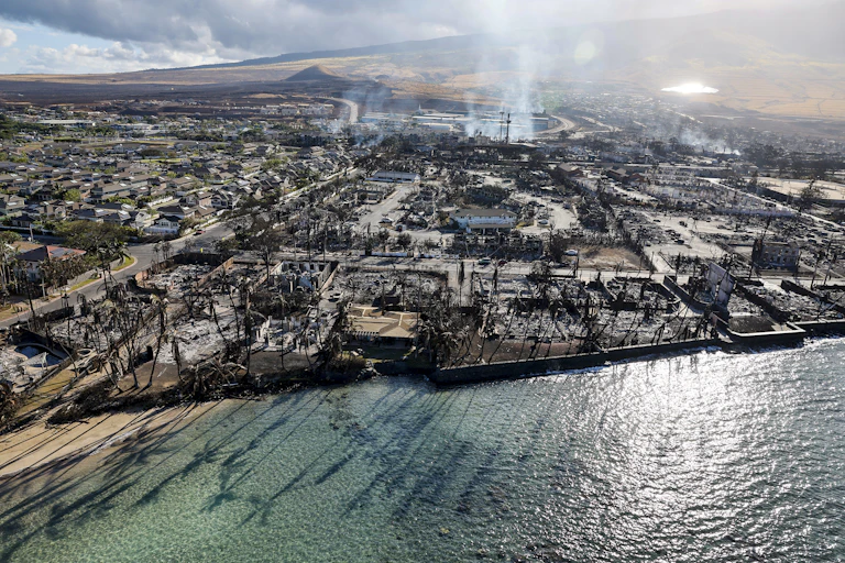 An aerial view from over the water toward town with burned buildings across the entire landscape and smoke rising beyond.