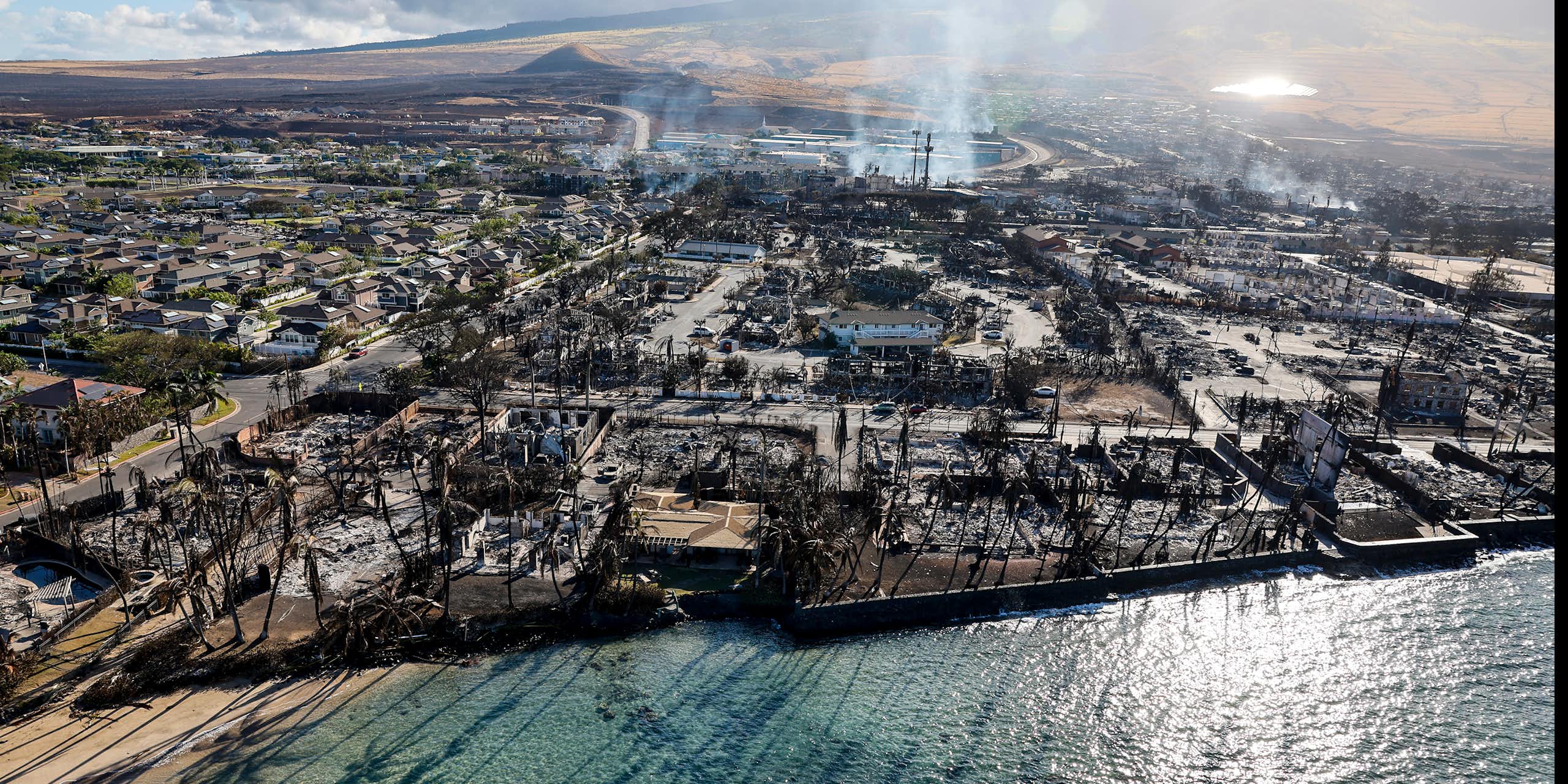 An aerial view from over the water toward town with burned buildings across the entire landscape and smoke rising beyond.