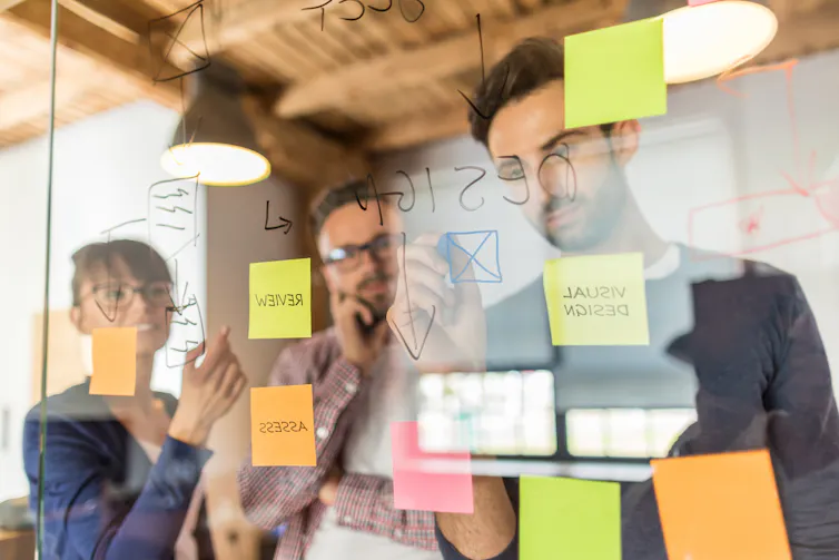 Three people stand behing a glass wall covered in sticky notes. One of the people is writing on the glass wall with a whiteboard marker.