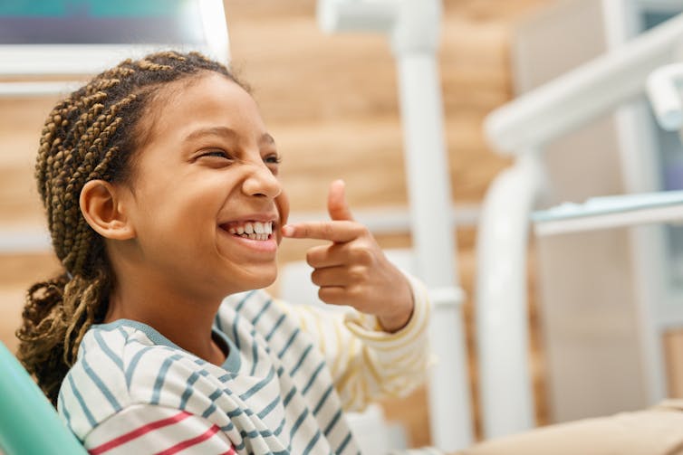 A girl smiles and points at her teeth.