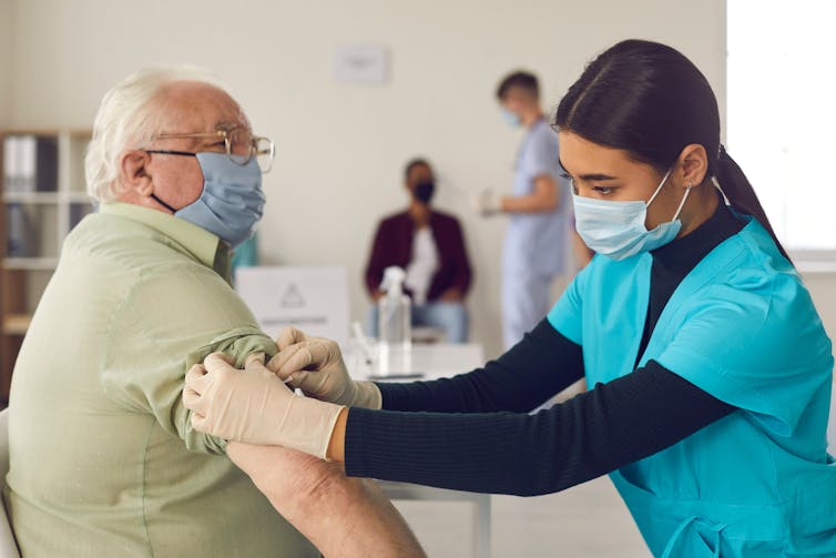 A nurse pushes up the left sleeve of an older man. Both are wearing masks.
