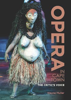 A woman on a stage wearing a grass skirt and a giant mask over her head complete with a prosthetic torso, her hands at her sides.