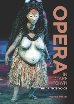 A woman on a stage wearing a grass skirt and a giant mask over her head complete with a prosthetic torso, her hands at her sides.