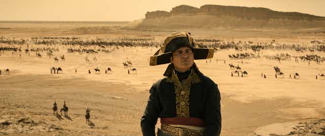 o ator Joaquin Phoenix caracterizado como Napoleão num deserto com camelos passando ao fundo. 
